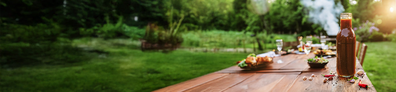 Glass jar with BBQ sauce, next to spices on a picnic table, with a barbeque in the background.