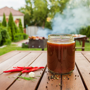 Glass jar with BBQ sauce, next to spices on a picnic table, with a barbeque in the background.