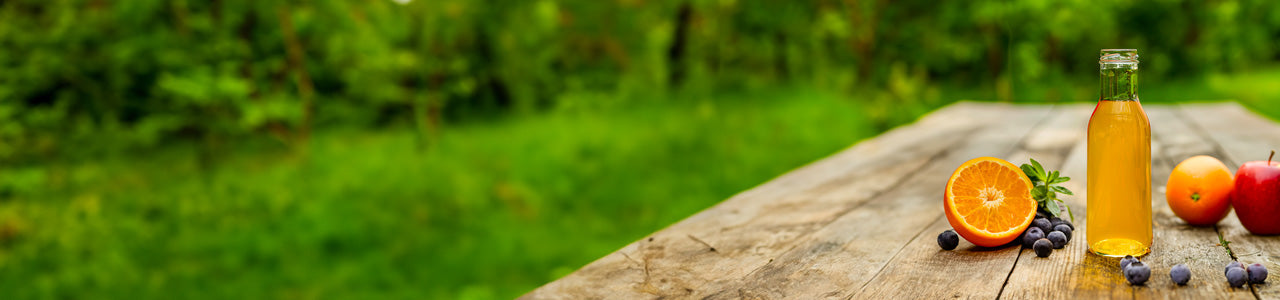 Glass bottle of juice on a wooden table with fruits and a blurred green summer background.